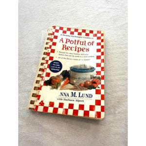 Peut inclure: Un livre de cuisine à reliure spirale intitulé A Potful of Recipes par Anna M. Lund avec Barbara Alpert. La couverture présente une bordure à carreaux rouges et blancs, du texte sur les repas en mijoteuse et une mijoteuse avec des légumes.