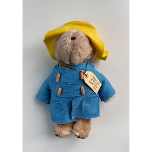 May include: A plush Paddington Bear toy wearing a blue coat with wooden toggles and a yellow hat. A tag attached to the coat reads "Please look after this BEAR. Thank You."