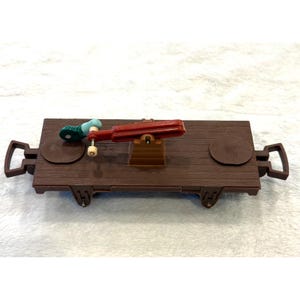 May include: A brown toy train car with a red and teal tool attached. The toy train car has two circular platforms and two handles. The tool is red with a teal and green handle.