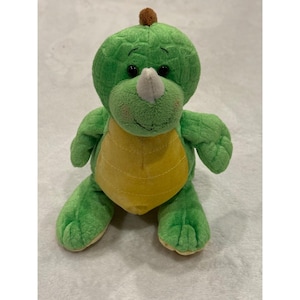 May include: A plush green dinosaur toy with a yellow belly and a white horn. The dinosaur has black eyes and a small brown bump on its head. The toy is sitting on a white surface.