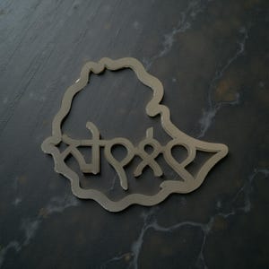 Puede incluir: Un contorno plateado del país de Etiopía, con escritura amárica en su interior. El objeto está sobre una superficie oscura y veteada. La escritura es una representación estilizada del nombre del país.