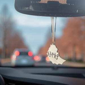 Puede incluir: Un ambientador de coche de madera con la forma de Etiopía, colgado de un espejo retrovisor. El ambientador tiene texto en amárico. El fondo muestra una escena de calle borrosa con coches y árboles.