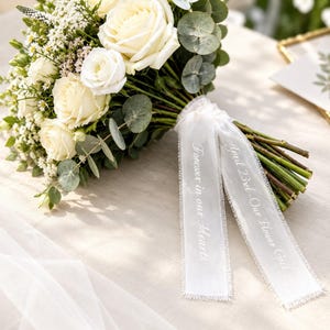 Op de afbeelding: Een bloemenboeket met witte rozen, kleine witte vulbloemen en groene eucalyptusbladeren. De stelen zijn omwikkeld met twee witte linten met gerafelde randen, bedrukt met de tekst "Forever in our Hearts" en "April 23rd - Our Flower Girl".