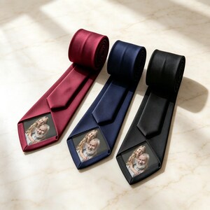 May include: Three neckties in burgundy, navy blue, and black, each featuring a smooth, satin-like finish. Each tie displays a rectangular photo print near the pointed tip, showing a portrait of a young girl and an older man with a white beard.