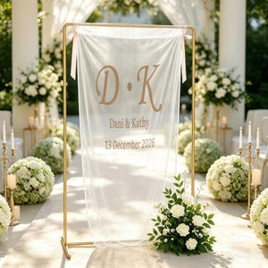 May include: A rectangular gold metal frame holding a sheer white fabric banner with light pink ribbon ties. The banner features the copper-coloured text D K, the names Dani & Kathy, and the date 13 December 2026 printed in a serif font.