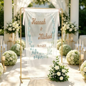 Può includere: Una cornice rettangolare in metallo dorato che sostiene uno stendardo in tessuto bianco trasparente con illustrazioni di foglie azzurre e il testo Hannah & Michael 16.11.2026 in lettere marroni. Lo stendardo è fissato con nastri rosa chiaro.