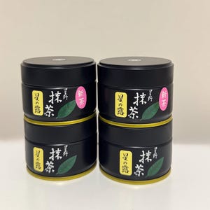 Könnte beinhalten: Vier zylindrische Teedosen aus schwarzem Metall, die in zwei Säulen gestapelt sind. Jede Dose hat ein gelbes Etikett mit japanischem Text, eine grüne Blattillustration und einen kleinen rosa Aufkleber mit japanischem Text oben.