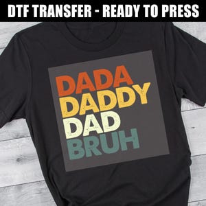 May include: Black t-shirt with a colorful graphic that reads "DADA DADDY DAD BRUH" in orange, yellow, white, and teal lettering. The words are stacked on top of each other. The words are inside a dark gray square.