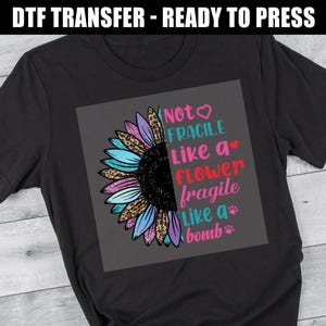 May include: Black t-shirt featuring a colorful sunflower design with the text "Not Fragile Like a Flower, Fragile Like a Bomb." The sunflower has blue, purple, and leopard print petals. The text is in pink, blue, and red.