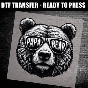 May include: Black and white DTF transfer featuring a detailed illustration of a bear wearing sunglasses. The sunglasses display the words "PAPA BEAR". The image is set against a gray background and a brick wall.