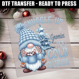 May include: DTF transfer with a winter gnome design. The gnome wears a blue hat and mittens, holding a mug of hot cocoa. Text reads "Snuggle Up, Gnomies, It's Cold Outside." The image has a winter theme with snowflakes and a Christmas tree.