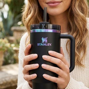 Puede incluir: Un vaso Stanley negro con asa y pajita. El vaso tiene una tapa negra con detalles iridiscentes y el logotipo de Stanley. La persona sostiene el vaso.