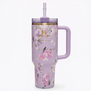 May include: A lavender Stanley tumbler with a floral design featuring pink and purple flowers, a bird, and gold trim. The tumbler has a handle, a lid, and a straw. The Stanley logo is visible on the front.