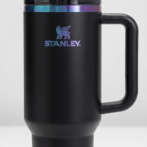 Puede incluir: Vaso Stanley negro con asa y pajita. El vaso tiene una tapa negra y una banda iridiscente alrededor de la parte superior. El logotipo de Stanley est&aacute; impreso en colores iridiscentes en la parte delantera del vaso.