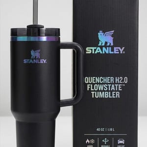 Puede incluir: Vaso Stanley Quencher H2.0 Flowstate negro con asa y pajita reutilizable. El vaso tiene una capacidad de 1,18 L y es compatible con portavasos de coche. El logotipo de Stanley es visible.