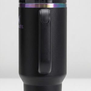 Puede incluir: Vaso t&eacute;rmico negro con asa y pajita. El vaso tiene una tapa negra con una banda iridiscente arco&iacute;ris. El logotipo de Stanley es visible. Dise&ntilde;ado para mantener las bebidas fr&iacute;as o calientes.