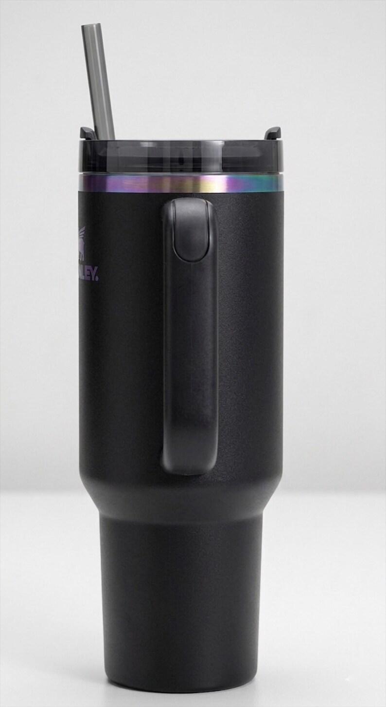 Puede incluir: Vaso t&eacute;rmico negro con asa y pajita. El vaso tiene una tapa negra con una banda iridiscente arco&iacute;ris. El logotipo de Stanley es visible. Dise&ntilde;ado para mantener las bebidas fr&iacute;as o calientes.