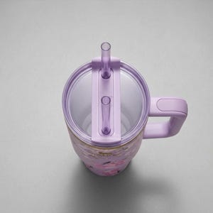 May include: A floral-patterned tumbler with a lavender lid, handle, and straw. The tumbler features a clear, transparent lid with a straw and a handle for easy carrying. The floral design includes pink and gold accents.
