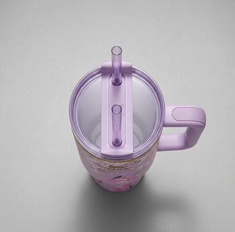 May include: A floral-patterned tumbler with a lavender lid, handle, and straw. The tumbler features a clear, transparent lid with a straw and a handle for easy carrying. The floral design includes pink and gold accents.