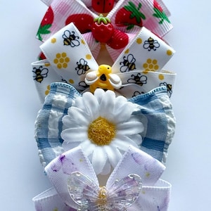 May include: A selection of decorative hair bows with diverse designs. The bows feature ladybirds, strawberries, bees, daisies, and butterflies. The bows are made of ribbon and adorned with embellishments, ideal for adding a touch of whimsy to any hairstyle.