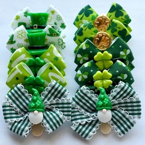 May include: A collection of green and white St. Patrick's Day hair bows. The bows feature various designs, including shamrocks, leprechaun hats, and gnome figures. The bows are made of ribbon and adorned with decorative elements, perfect for festive celebrations.