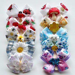 May include: A collection of colorful hair bows with various designs. The bows feature ladybugs, strawberries, bees, daisies, clouds, butterflies, and mushrooms. Each bow has a unique embellishment, adding to their charm. These accessories are perfect for adding a touch of whimsy to any hairstyle.