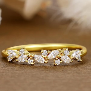 May include: A gold ring with a floral design. The ring features small, clear, marquise-cut and round-cut stones set in a delicate pattern. The band is a thin, polished gold. The ring is displayed on a brown surface.