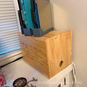 May include: A wooden storage box with a series of slots on the front, holding several file folders in a gray plastic holder. The folders are black, teal, and tan. The box is sitting on a white surface.