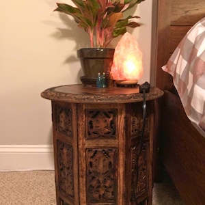May include: A dark brown, carved wooden side table with a round top. The table has intricate floral designs and holds a potted plant, a glowing Himalayan salt lamp, and small glass bottles. The table is next to a bed.