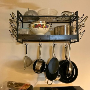 May include: A black metal wall-mounted pot rack with a wooden shelf, featuring a wire grid top for lids and containers, and multiple hooks underneath for hanging various stainless steel and non-stick pans, a colander, and a small mesh strainer.