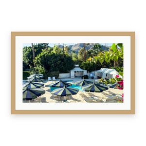 May include: A framed photograph of a sunny outdoor pool area featuring multiple dark blue and green striped patio umbrellas, lounge chairs, white cabanas, and lush green trees set against a backdrop of mountains under a clear blue sky.