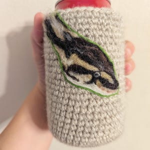 May include: A crocheted beige can cooler with a green, brown and black snake design.