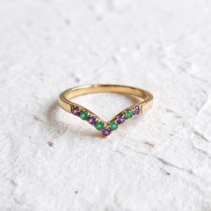 May include: A gold V-shaped ring featuring a row of alternating round-cut green and purple gemstones set along the chevron-style band. The metal has a polished finish, and the stones are arranged in a symmetrical pattern across the front.