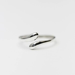 May include: A silver-colored ring with a unique design. The ring features two teardrop-shaped ends that do not fully connect, creating an open, adjustable style. The ring is set against a white background.