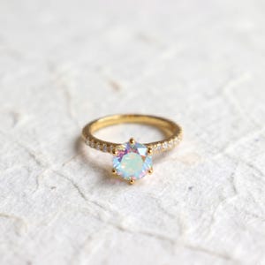 Puede incluir: Un anillo de oro con una gran gema hexagonal iridiscente en el centro. La banda también es dorada e incrustada con pequeñas gemas transparentes. El anillo se encuentra sobre un fondo texturizado blanquecino.