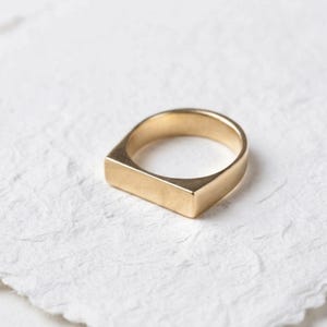 Op de afbeelding: Een goudkleurige ring met een glad, plat, rechthoekig balkje aan de bovenzijde. De ringband is massief en doorlopend, met een gepolijste metalen afwerking over het gehele sieraad. Het ontwerp is eenvoudig, met strakke lijnen en een uniforme, solide constructie.