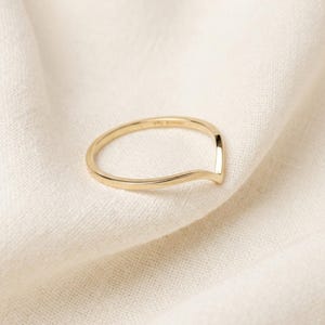 May include: A gold-colored ring with a V-shaped design. The ring is set against a white fabric background. The ring's band is thin and smooth, with a subtle curve. The ring is likely made of gold or a gold-colored metal.