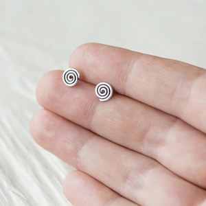 May include: A pair of small, silver-colored stud earrings. Each earring features a spiral design with a dark, contrasting center. The earrings are displayed on a light background, showcasing their minimalist and modern aesthetic.