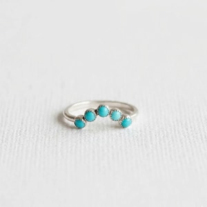 May include: A silver ring with five turquoise-colored stones set in a curved design. The ring is set against a white background, highlighting the bright blue of the stones.