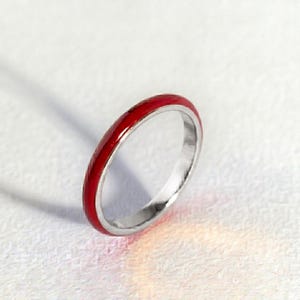 May include: A thin, circular ring featuring a smooth, vibrant red enamel inlay set within a polished silver-toned metal band. The band has a rounded profile and a consistent width, showcasing a clean, simple design with a reflective metallic finish.