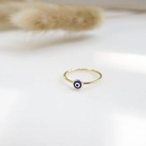 May include: A gold ring with a small, circular charm displaying a blue eye design. The ring has a slender band and a minimalist style. The eye charm has a white centre, a blue iris, and a black outline.