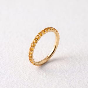Op de afbeelding: Een dunne gouden ring met een doorlopende rij kleine, ronde, felgele edelstenen die langs de buitenomtrek zijn gezet. Het metaal heeft een gepolijste afwerking en de stenen zijn gelijkmatig verdeeld voor een uniform ontwerp.