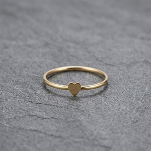 May include: A delicate gold ring with a small heart-shaped charm. The ring has a thin band and a polished finish, creating a minimalist and elegant aesthetic. The heart charm is centered on the band.