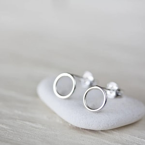 May include: A pair of silver-colored, open-circle stud earrings. The earrings are displayed on a white, smooth, oval-shaped stone. The earrings have a simple, minimalist design, with a circular frame and a hollow center. The background is a soft, blurred white.