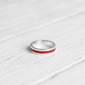 May include: A silver band ring featuring a smooth, polished finish with a single, vibrant red horizontal stripe inlay that runs continuously around the centre of the outer surface, creating a clean and simple aesthetic for this piece of jewellery.