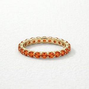 Könnte beinhalten: Ein goldener Eternity-Ring mit einer durchgehenden Reihe rund geschliffener, leuchtend orangefarbener Edelsteine in einer polierten Metallfassung. Der Ring zeigt ein gleichmäßiges kreisförmiges Design, bei dem jeder Stein von kleinen Metallkrappen gehalten wird.