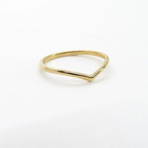 Puede incluir: Un anillo dorado con un diseño en forma de V. El anillo está hecho de un metal brillante y liso y se encuentra sobre una superficie blanca. El diseño del anillo es simple y elegante, adecuado para el uso diario o ocasiones especiales.