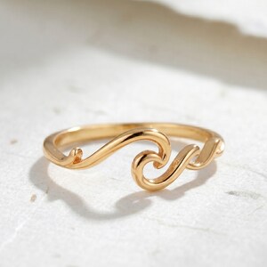 May include: A gold ring with a wave design. The ring has a flowing, curved shape, resembling a stylized ocean wave. The ring is made of shiny, polished metal and is set against a neutral background.