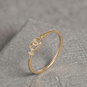 May include: A gold-colored ring featuring a double female symbol design. The ring is thin and delicate, with the symbols connected at the center and at the top and bottom of the ring.