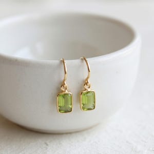May include: A pair of gold drop earrings featuring rectangular, light green faceted gemstones. Each earring is suspended from a simple gold hook, showcasing a clear, vibrant green stone set in a polished gold frame that catches the light.
